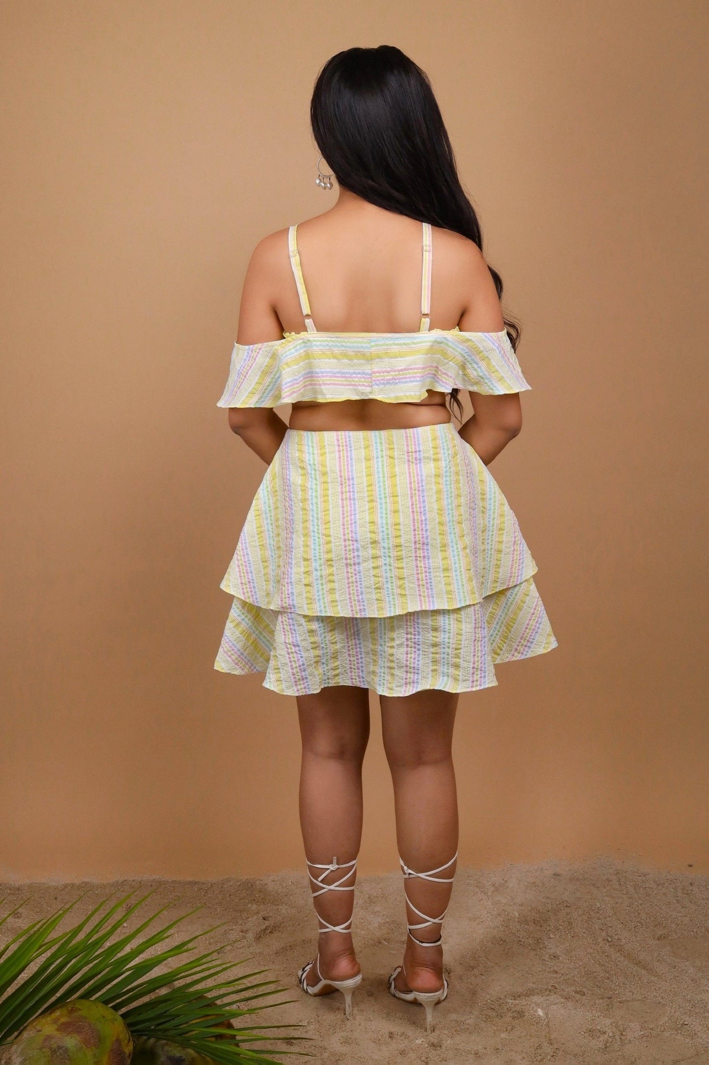 Woman sitting on a wicker chair holding a coconut against a beige background wearing an off-shoulder yellow color striped dress by Soliesse. Its a perfect dress for a sunny day, brunch, date, a vacation or getaway. Its chic & comfortable at the same time.