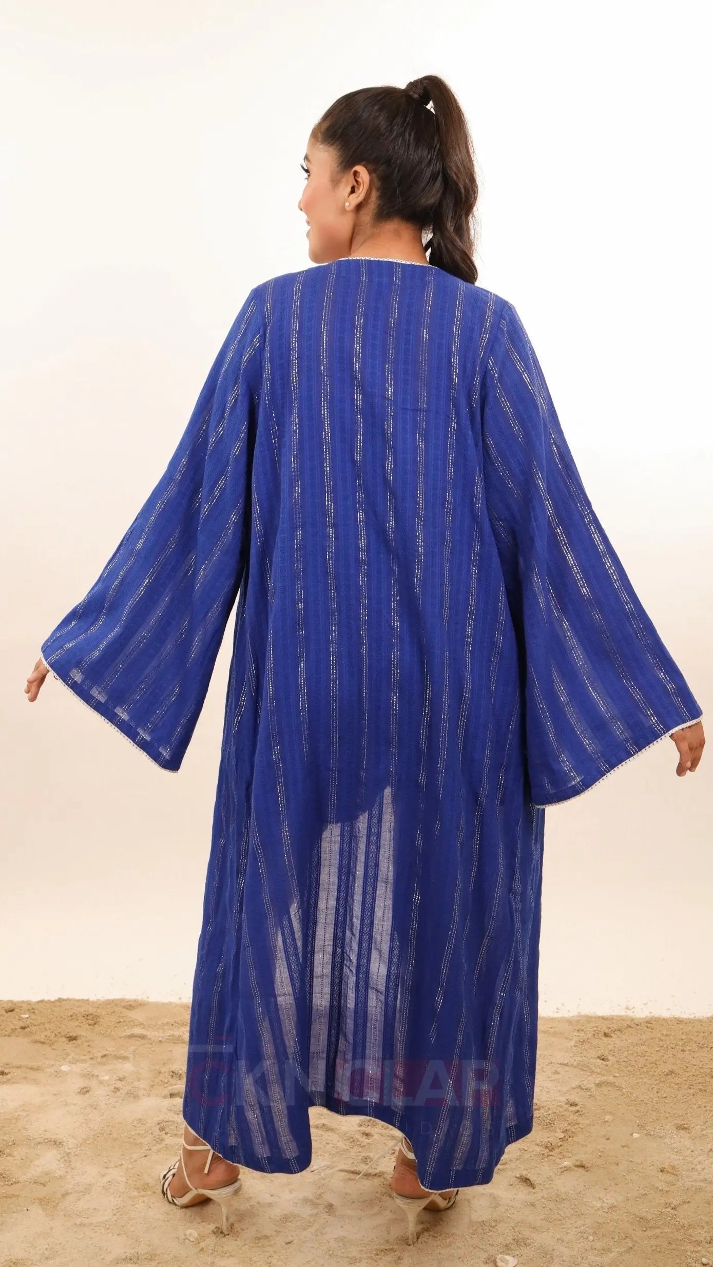 Woman wearing Blue color shrug or open kimono you could say called Nova Kimono by Soliesse brand. Its an open shrug style midi length, fabric is lurex cotton dobby in striped pattern vertical, contrast white pom pom lace detailing, bell sleeves. Can be styled multiple ways, perfect for vacations, getaway or regular summer days or outings.