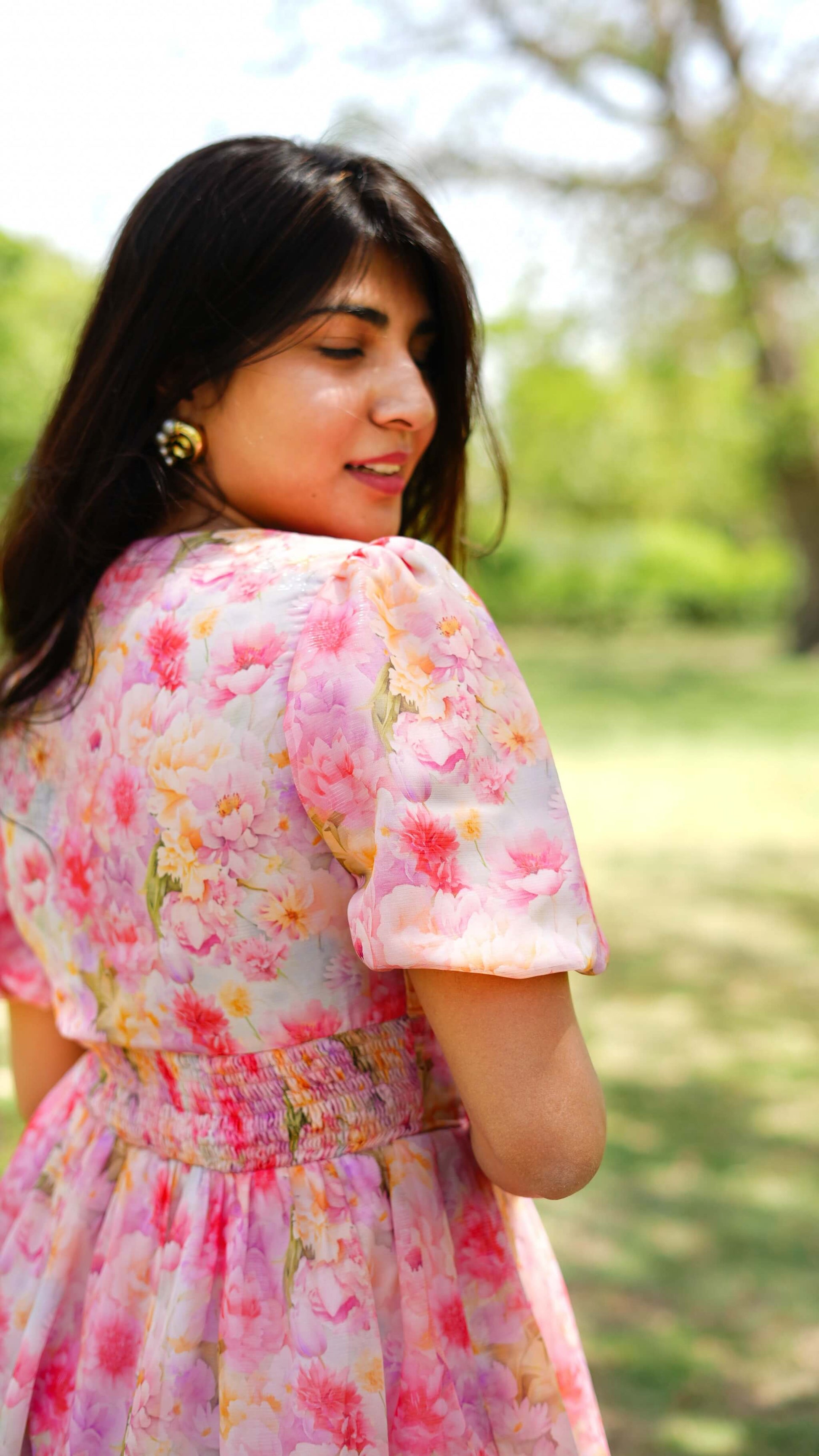 A woman wearing a pink floral dress with puff sleeves, shown from behind in an outdoor park setting with green trees in the background.