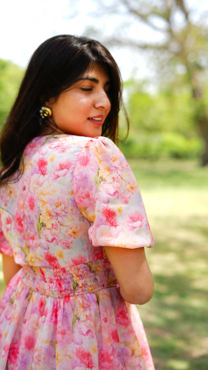 A woman wearing a pink floral dress with puff sleeves, shown from behind in an outdoor park setting with green trees in the background.