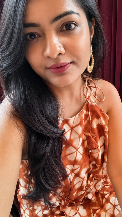 A close-up portrait of a woman wearing a brown and white tie-dye patterned halter dress.. gold hoop earrings, set against a dark red curtain background.
