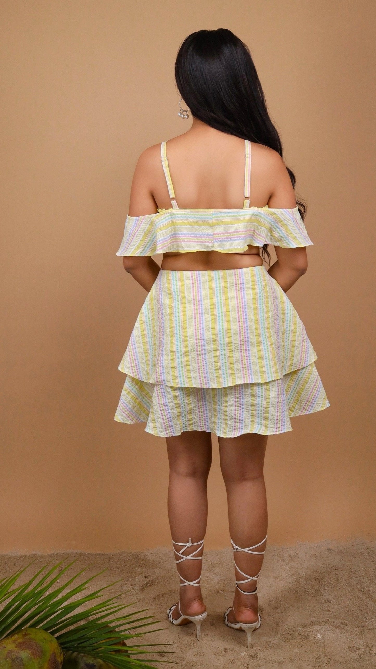 Woman sitting on a wicker chair holding a coconut against a beige background wearing an off-shoulder yellow color striped dress by Soliesse. Its a perfect dress for a sunny day, brunch, date, a vacation or getaway. Its chic & comfortable at the same time.