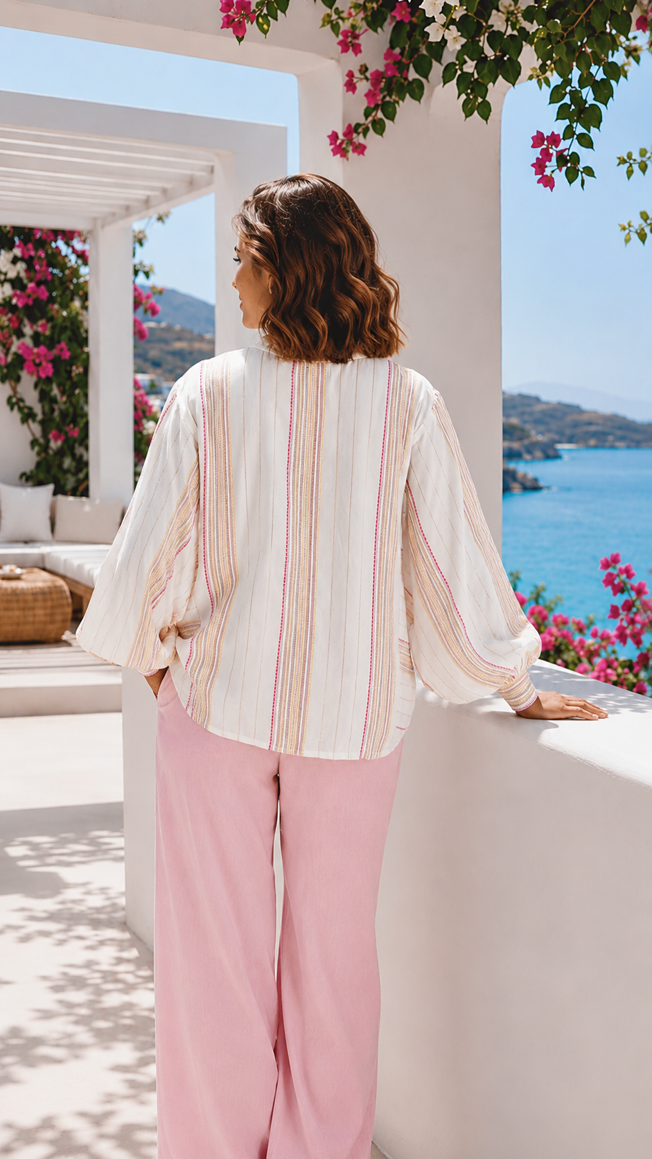 Back view of the Woman in a striped shirt and pink pants standing on a balcony with a scenic view of the sea.
